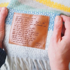 May include: A light gray wool blanket featuring a rectangular brown leather patch stitched onto the fabric. The patch is engraved with Shakespeare's Sonnet 116 in cursive script. The blanket has horizontal stripes in yellow, teal, and blue.