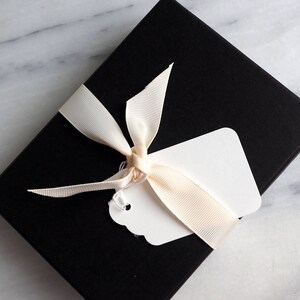 K&ouml;nnte beinhalten: Eine schwarze Geschenkbox mit einem wei&szlig;en Etikett und einem cremefarbenen Band, das zu einer Schleife gebunden ist.