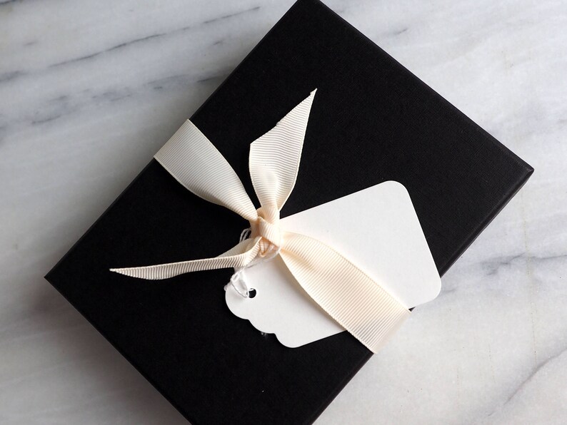 K&ouml;nnte beinhalten: Eine schwarze Geschenkbox mit einem wei&szlig;en Etikett und einem cremefarbenen Band, das zu einer Schleife gebunden ist.