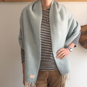 May include: A light blue herringbone patterned scarf with white fringe. The scarf is draped around a person wearing a striped shirt and brown pants.