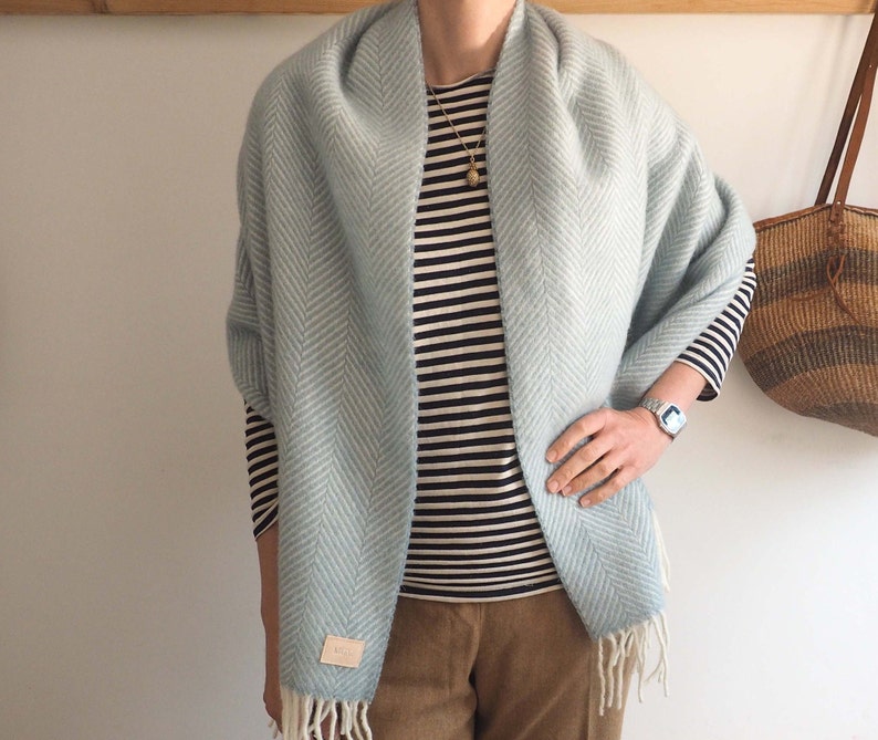 May include: A light blue herringbone patterned scarf with white fringe. The scarf is draped around a person wearing a striped shirt and brown pants.
