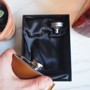 K&ouml;nnte beinhalten: Eine braune Leder-H&uuml;ftflasche mit silbernem Verschluss und einem silbernen Trichter in einer schwarzen, mit Samt ausgekleideten Box.