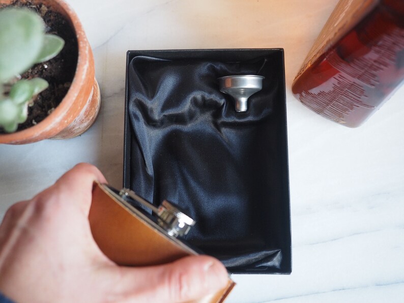 K&ouml;nnte beinhalten: Eine braune Leder-H&uuml;ftflasche mit silbernem Verschluss und einem silbernen Trichter in einer schwarzen, mit Samt ausgekleideten Box.