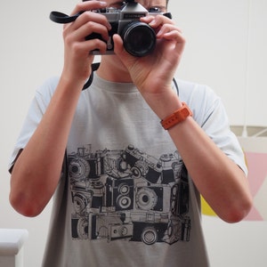 May include: A grey t-shirt with a black and white print of vintage cameras. The shirt features a variety of camera styles, including a rangefinder, a single-lens reflex, and a box camera.