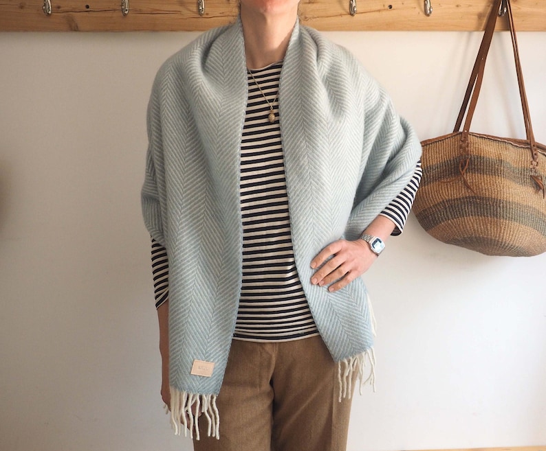 May include: A light blue herringbone patterned scarf with fringe. The scarf is draped around a person wearing a striped shirt and brown pants.