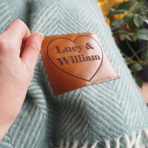 May include: A light green and white herringbone patterned throw blanket featuring a brown leather patch. The patch is heart-shaped and engraved with the text Lucy & William in a dark font, centered on the textured fabric of the blanket.