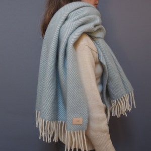 May include: A light blue herringbone wool scarf with white fringe. The scarf is draped around a person's neck and shoulders.