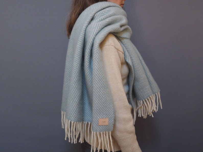 May include: A light blue herringbone wool scarf with white fringe. The scarf is draped around a person's neck and shoulders.