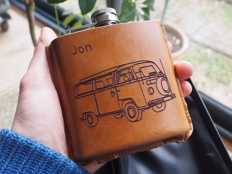 K&ouml;nnte beinhalten: Eine braune Lederflasche mit silbernem Verschluss und einer personalisierten Gravur mit dem Text "Jon". Die Flasche zeigt ein eingegraveertes Design eines alten Volkswagen-Transporters.