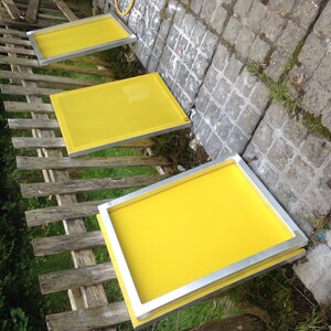May include: Three rectangular screen printing frames with bright yellow mesh stretched across silver metal borders. The frames are arranged in a diagonal line, showcasing the smooth, taut surface of the yellow fabric against the metallic frames.