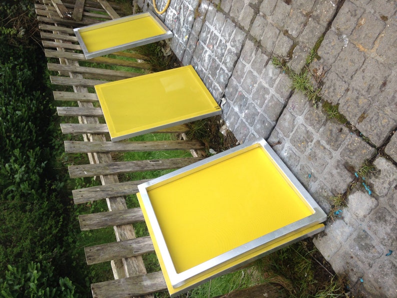 May include: Three rectangular screen printing frames with bright yellow mesh stretched across silver metal borders. The frames are arranged in a diagonal line, showcasing the smooth, taut surface of the yellow fabric against the metallic frames.