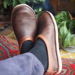 Könnte beinhalten: Ein Paar braune Leder-Slipper mit hellbraunem Besatz und weißen Sohlen. Die Schuhe werden mit schwarzen Socken und einer blauen Hose getragen. Die Schuhe stehen auf einem farbenfrohen gemusterten Teppich.