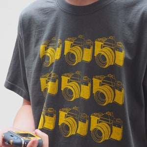 May include: A charcoal gray short-sleeve t-shirt featuring a repeating grid pattern of nine yellow graphic illustrations of vintage SLR cameras. The design is screen-printed on the front of the shirt, showcasing detailed camera lenses and bodies.