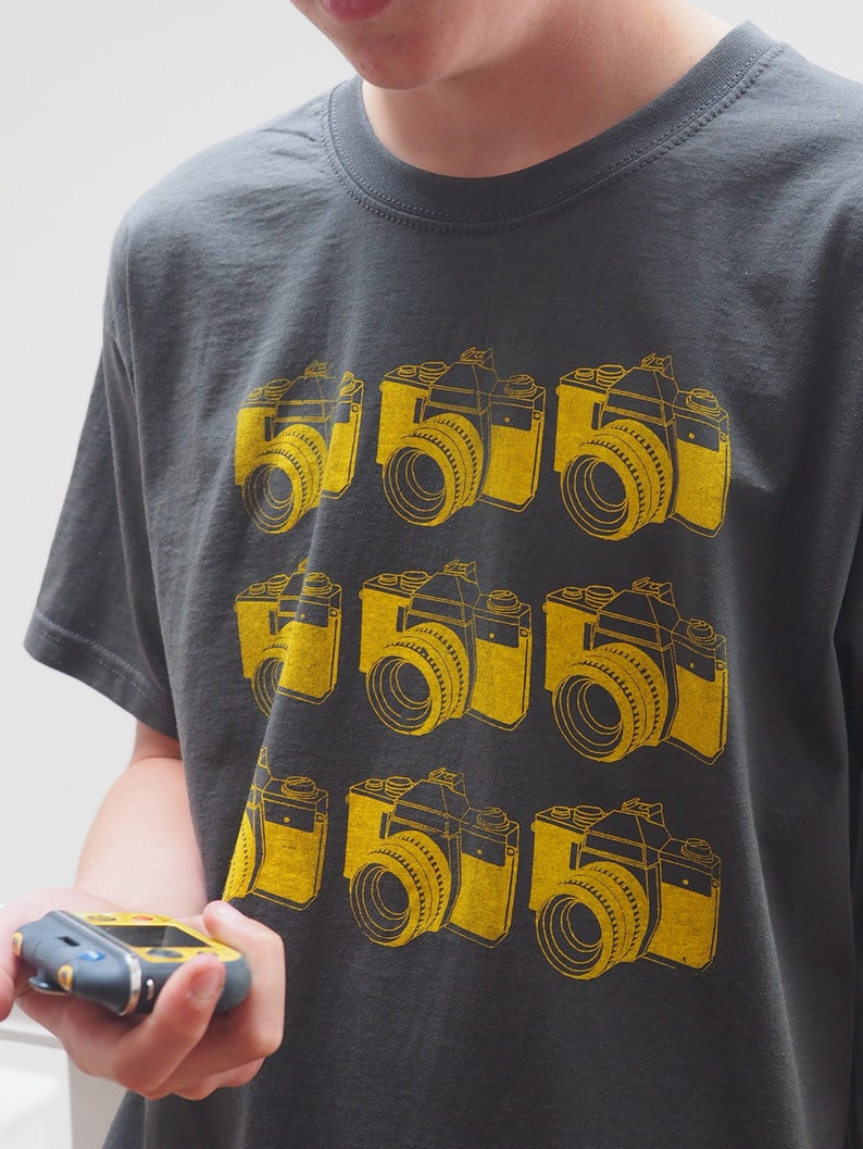 May include: A charcoal gray short-sleeve t-shirt featuring a repeating grid pattern of nine yellow graphic illustrations of vintage SLR cameras. The design is screen-printed on the front of the shirt, showcasing detailed camera lenses and bodies.
