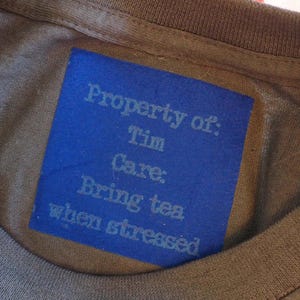 May include: A brown t-shirt featuring a rectangular blue label sewn onto the inner neckline. The label displays the text: Property of: Tim Care: Bring tea when stressed, printed in a light blue font against the solid blue background.