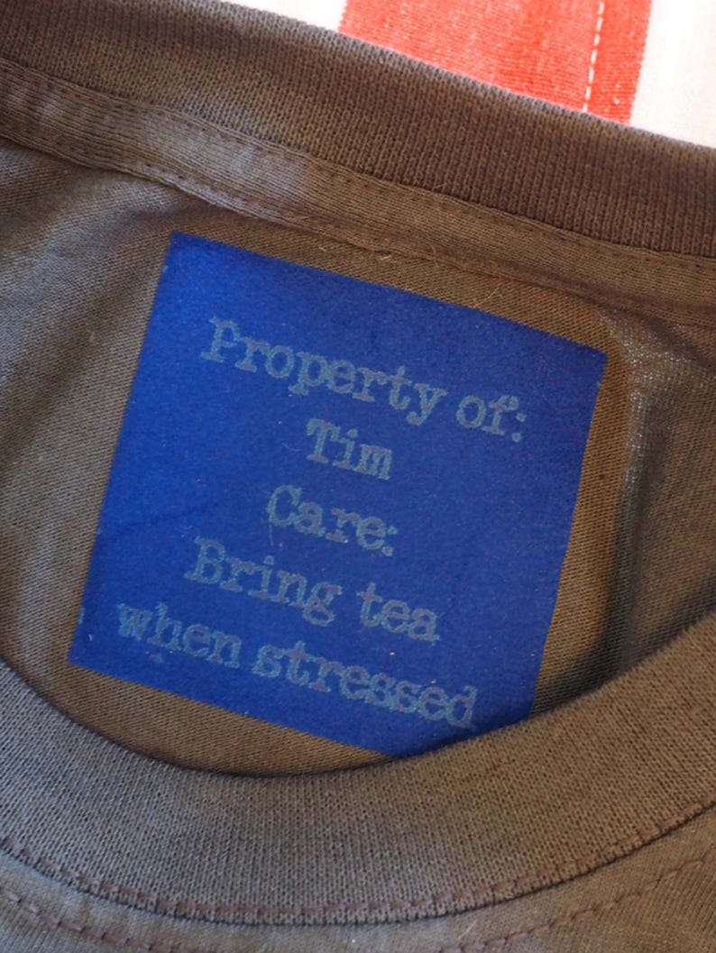 May include: A brown t-shirt featuring a rectangular blue label sewn onto the inner neckline. The label displays the text: Property of: Tim Care: Bring tea when stressed, printed in a light blue font against the solid blue background.
