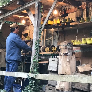 May include: A cobbler working in a rustic outdoor workshop. The man wears a blue jacket and cap, focused on his craft. Shelves display shoe lasts in yellow and green. The workshop is illuminated by hanging light bulbs, with tools and materials visible.