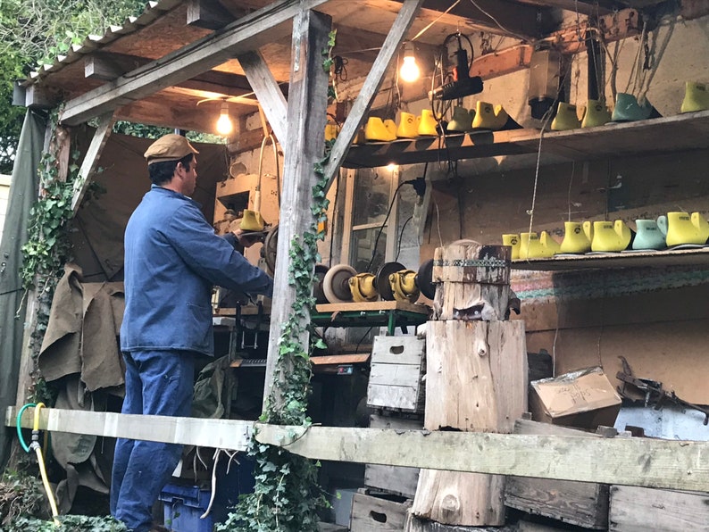 May include: A cobbler working in a rustic outdoor workshop. The man wears a blue jacket and cap, focused on his craft. Shelves display shoe lasts in yellow and green. The workshop is illuminated by hanging light bulbs, with tools and materials visible.