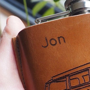 K&ouml;nnte beinhalten: Eine braune Lederflasche mit silbernem Verschluss und dem eingravierten Namen "Jon" auf der Vorderseite.