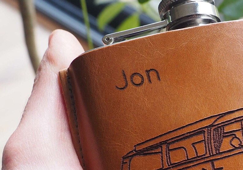 K&ouml;nnte beinhalten: Eine braune Lederflasche mit silbernem Verschluss und dem eingravierten Namen "Jon" auf der Vorderseite.
