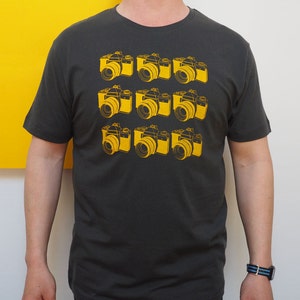 May include: A charcoal gray short-sleeve t-shirt featuring a repeating grid pattern of nine yellow line-art illustrations of vintage film cameras arranged in three rows of three, centered on the front of the garment.