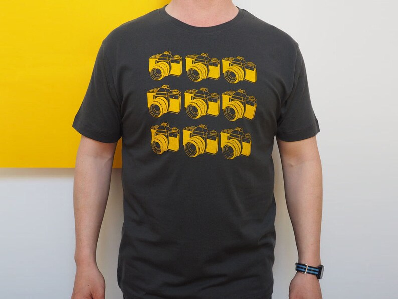 May include: A charcoal gray short-sleeve t-shirt featuring a repeating grid pattern of nine yellow line-art illustrations of vintage film cameras arranged in three rows of three, centered on the front of the garment.