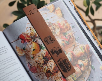 May include: A rectangular brown leather bookmark featuring the engraved letters JLS at the top, followed by three distinct black illustrations of vintage cameras arranged vertically along the centre of the smooth surface.