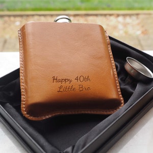 K&ouml;nnte beinhalten: Eine braune Leder-Taschenflasche mit silbernem Verschluss und einem Trichter. Die Flasche ist mit dem Text "Happy 40th Little Bro" graviert.