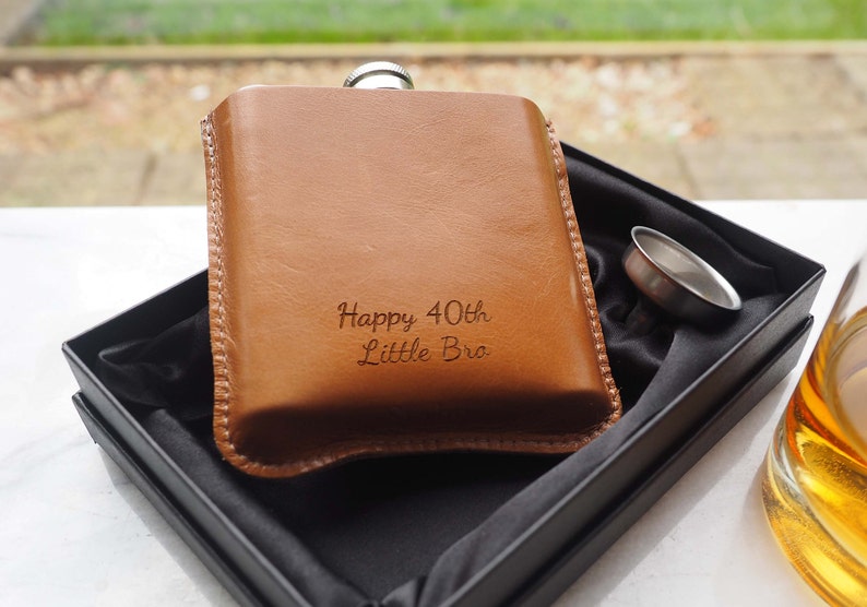 K&ouml;nnte beinhalten: Eine braune Leder-Taschenflasche mit silbernem Verschluss und einem Trichter. Die Flasche ist mit dem Text "Happy 40th Little Bro" graviert.