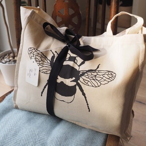 May include: A white canvas tote bag with black handles and a black ribbon tied around it. The bag has a black and white illustration of a bumblebee on the front. The bag has a white tag attached to it that says "The Little Bee Shop".