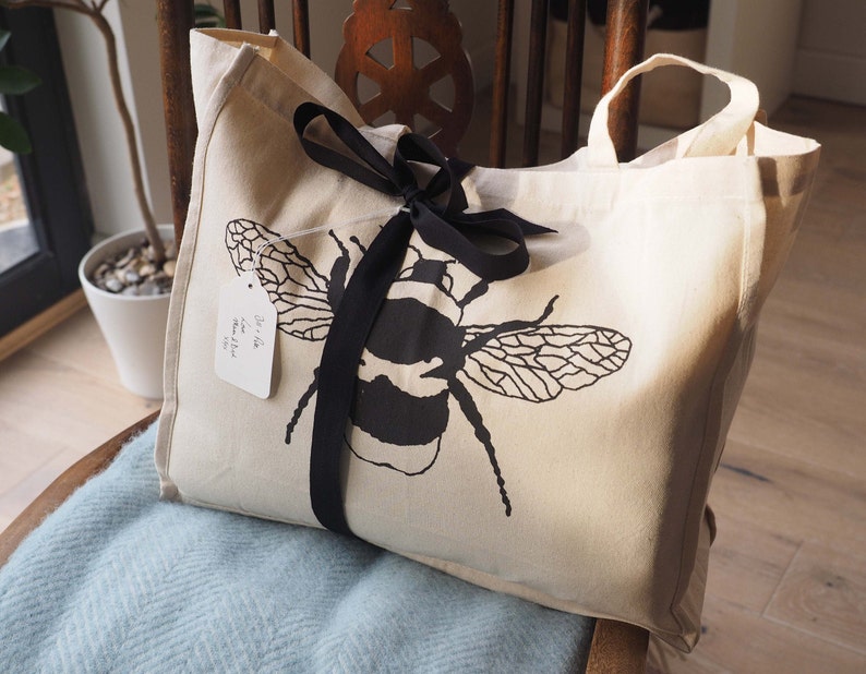 May include: A white canvas tote bag with black handles and a black ribbon tied around it. The bag has a black and white illustration of a bumblebee on the front. The bag has a white tag attached to it that says "The Little Bee Shop".