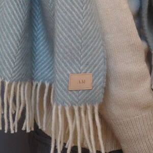 May include: A light blue and white herringbone patterned scarf with fringe. The scarf has a brown leather tag with the initials "AM" on it.