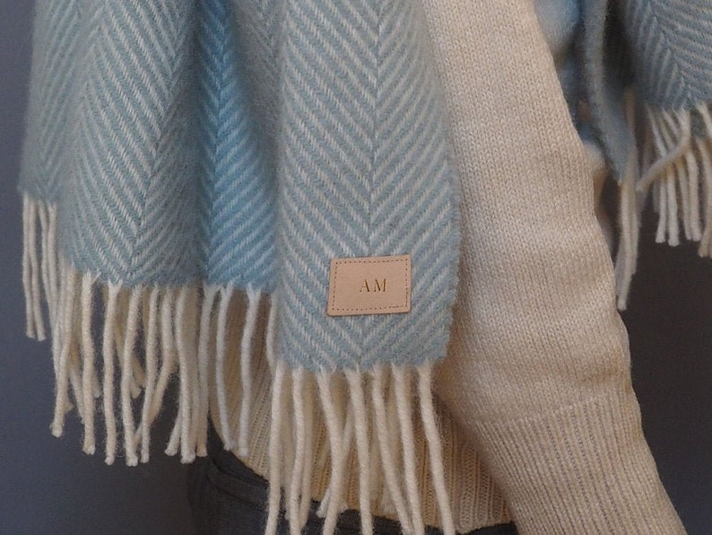 May include: A light blue and white herringbone patterned scarf with fringe. The scarf has a brown leather tag with the initials "AM" on it.