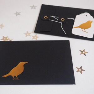 May include: Two black envelopes featuring a gold bird silhouette. One envelope is closed with white string wrapped around two black eyelets and has a white tag with a gold bird illustration attached. Small gold star-shaped confetti pieces are scattered.
