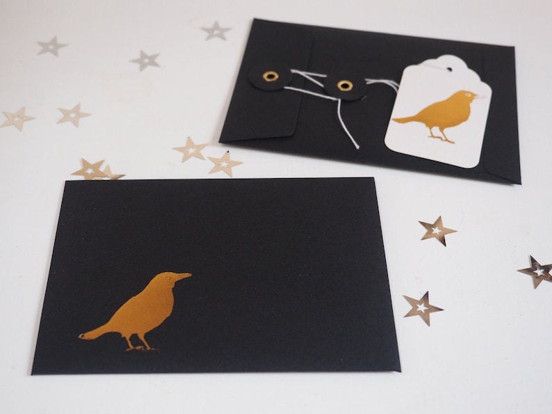 May include: Two black envelopes featuring a gold bird silhouette. One envelope is closed with white string wrapped around two black eyelets and has a white tag with a gold bird illustration attached. Small gold star-shaped confetti pieces are scattered.