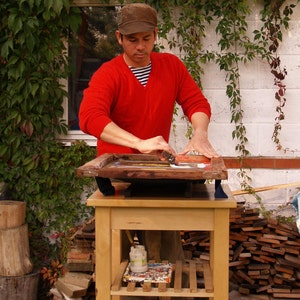 May include: A person wearing a red V-neck sweater and a brown cap uses a squeegee to apply white ink to a wooden screen printing frame. The frame rests on a light-colored wooden table with a lower shelf containing a small jar of white paint.