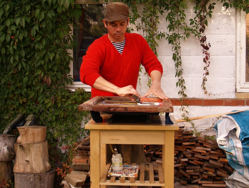 May include: A person wearing a red V-neck sweater and a brown cap uses a squeegee to apply white ink to a wooden screen printing frame. The frame rests on a light-colored wooden table with a lower shelf containing a small jar of white paint.