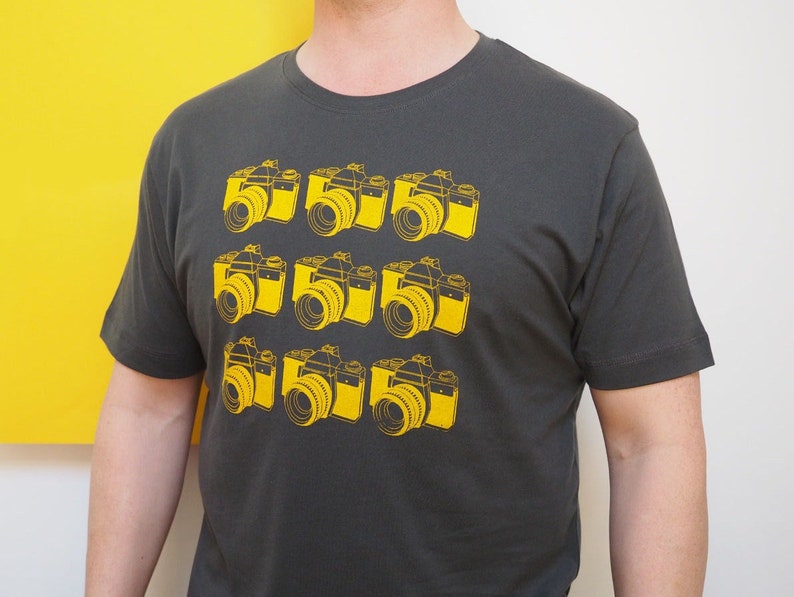 May include: A dark gray short-sleeved t-shirt featuring a repeating graphic print of nine yellow vintage-style cameras arranged in a three-by-three grid pattern across the front of the garment, with a crew neckline and a standard fit.