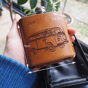 K&ouml;nnte beinhalten: Eine braune Lederflasche mit einer personalisierten Gravur "Jon" und einem eingegrabenen Bild eines Vintage-Vans. Die Flasche ist 11 cm hoch, 10 cm breit und 2,5 cm tief.