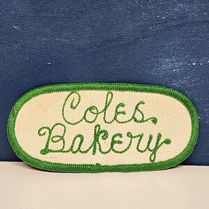 May include: A white oval-shaped patch with green embroidered edging. The text "Cole's Bakery" is embroidered in green.