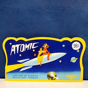 May include: A yellow and blue cardboard sign with a rocket ship and two people on it. The sign says "Atomic" and "98¢" and "Contains 85 assorted needles and 1 threader" and "Gold Eye".