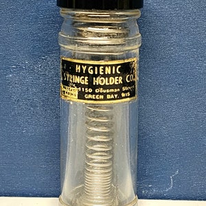 May include: A clear glass bottle with a black lid and a spring inside. The bottle has a label that reads "HYGIENIC SYRINGE HOLDER CO. 150 Ousman Street GREEN BAY, WIS."