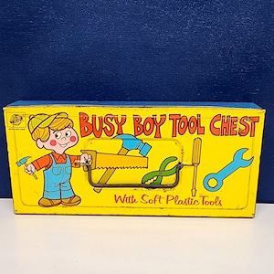 May include: A yellow metal toy tool chest with the words "Busy Boy Tool Chest" and "With Soft Plastic Tools" printed on the front. The chest features a cartoon boy holding a hammer and a variety of tools including a saw, pliers, a screwdriver, and a wrench.