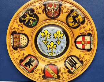 May include: A round wooden plate with a decorative design featuring coats of arms from different German cities. The plate has a central design of three stylized fleurs-de-lis in a blue and yellow color scheme. The text "Wiesbaden" is printed below the central design. The plate is decorated with a brown, gold, and white color scheme.