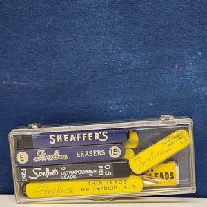May include: A clear plastic box filled with various writing implements, including a blue and yellow pencil with the text "Fineline" and "SHEAFFER'S" on it, a blue and yellow pencil with the text "Scripto" and "ULTRAPOLYMER LEADS" on it, and a blue and yellow pencil with the text "Fineline" and "THIN LEADS" on it.