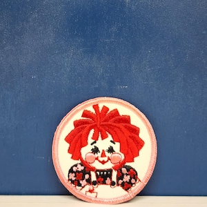 May include: Embroidered patch featuring a doll with red hair and rosy cheeks. The doll wears a black and red floral patterned dress. The patch has a white background with a pink border and is circular in shape. The patch is likely for clothing or accessories.