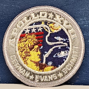 May include: A white embroidered patch with a silver border. The patch features a blue background with a white outline of a rocket ship, a yellow moon, and a red, white, and blue American flag. The text "Apollo 11" is written in a white circle around the image. The text "Edwin E. Aldrin Jr." is written in a white arc at the bottom of the patch.