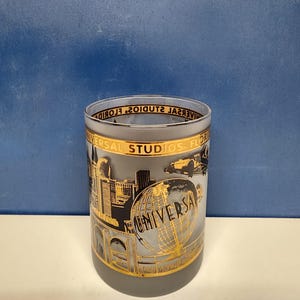 May include: A frosted glass featuring a gold and black design with the words "UNIVERSAL STUDIOS FLORIDA" and a globe graphic. The glass also depicts cityscapes and vintage vehicles.