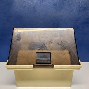 May include: A rectangular storage container with a solid off-white base and a hinged, translucent dark brown plastic lid. The lid features a central black rectangular latch mechanism with horizontal ridges for grip, allowing access to the interior.
