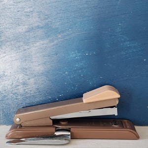 May include: A brown metal stapler with a silver staple remover. The stapler has a beige plastic top and a silver metal staple guide. The stapler is sitting on a blue surface.